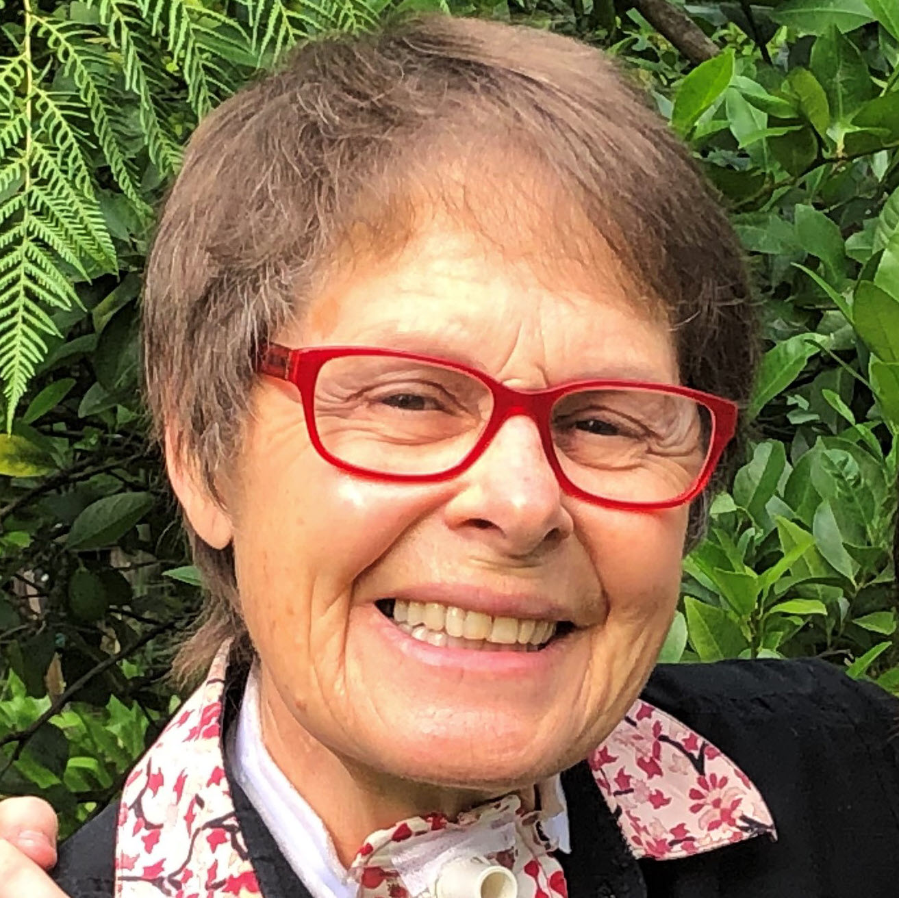 Denise Doraisamy smiling in red glasses with green foliage behind her