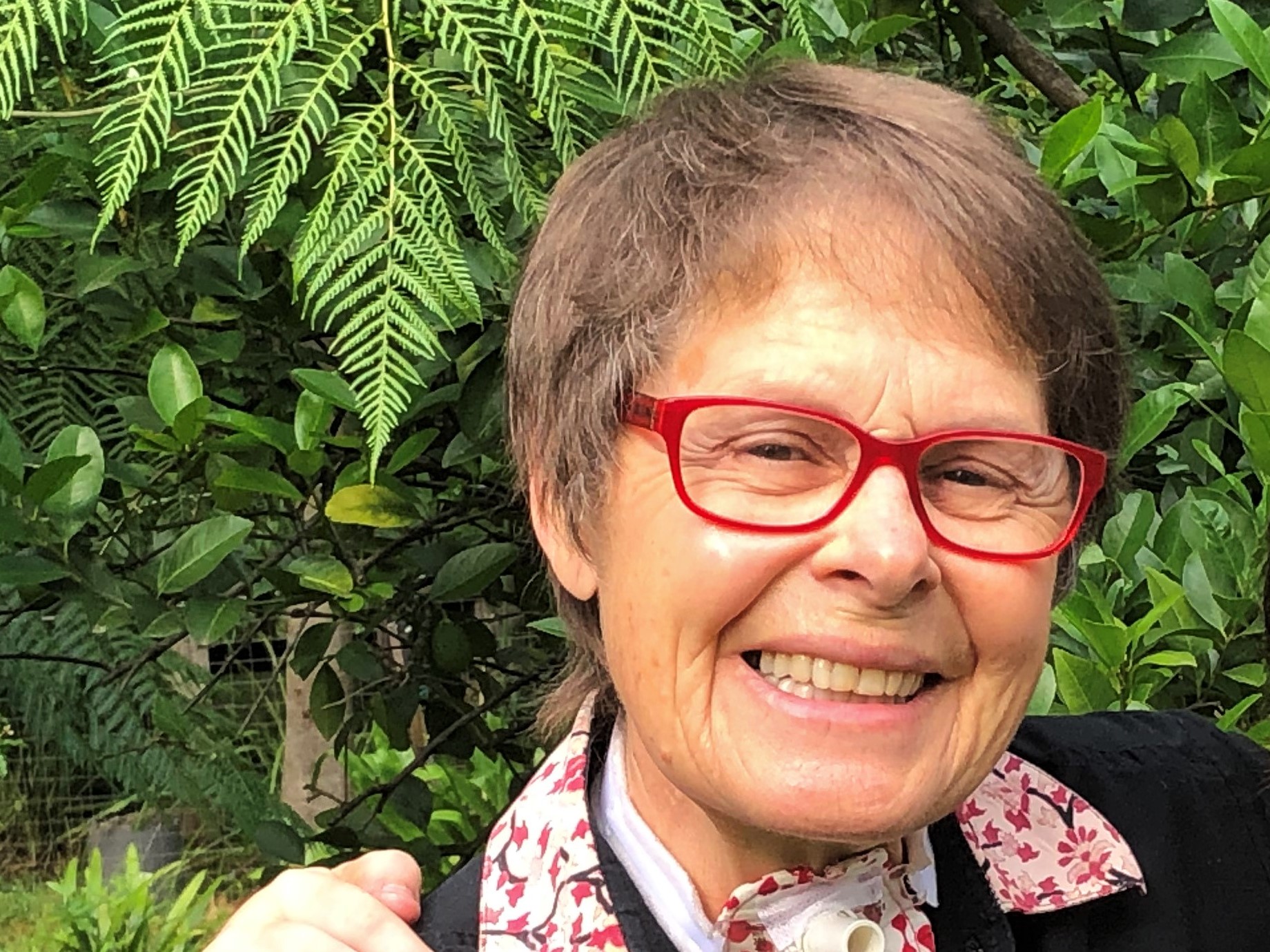 Portrait of Denise Doraisamy smiling outdoors in front of green foliage, wearing red glasses